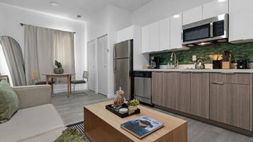 Eine moderne Küche mit weißen Schränken, einem Edelstahlkühlschrank und einer Mikrowelle. Es gibt einen Essbereich mit Tisch und Stühlen sowie ein Wohnzimmer mit Sofa und Couchtisch.
