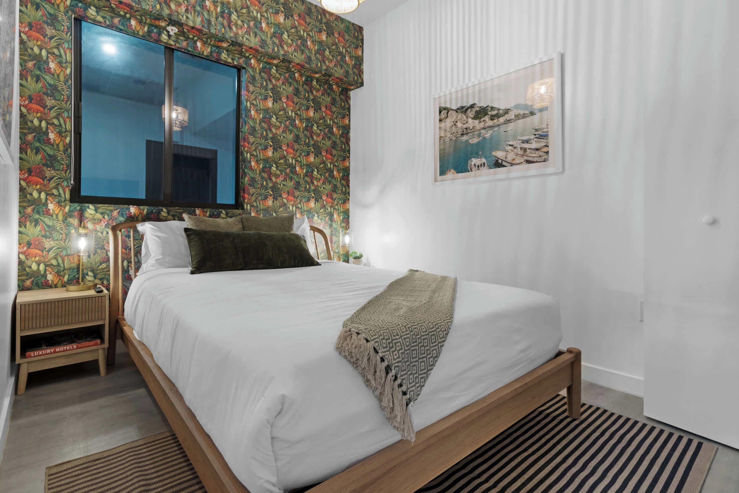 A bedroom with a large bed, a nightstand with a lamp, a window with a view, and a framed picture on the wall.