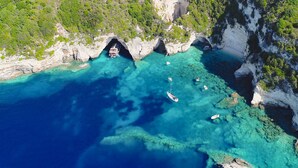 Vista aérea de una zona costera con aguas turquesas cristalinas, acantilados rocosos y una pequeña embarcación anclada cerca de la entrada de una cueva natural.