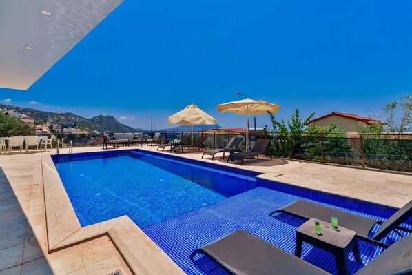 Piscina con sillas de descanso y sombrillas, rodeada de una terraza con baldosas y vistas a un paisaje montañoso.