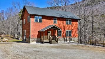 A two-story log cabin with a stone foundation, a porch, and a gravel driveway.