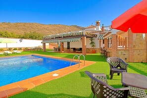 A swimming pool with a tiled edge, surrounded by artificial grass, a patio area with a table and chairs, and a building with a tiled roof and a red awning. - Experience authentic Costa del Sol living at Sara Sol, a beautiful 2 bedroom villa with private pool near Nerja (Nerja)