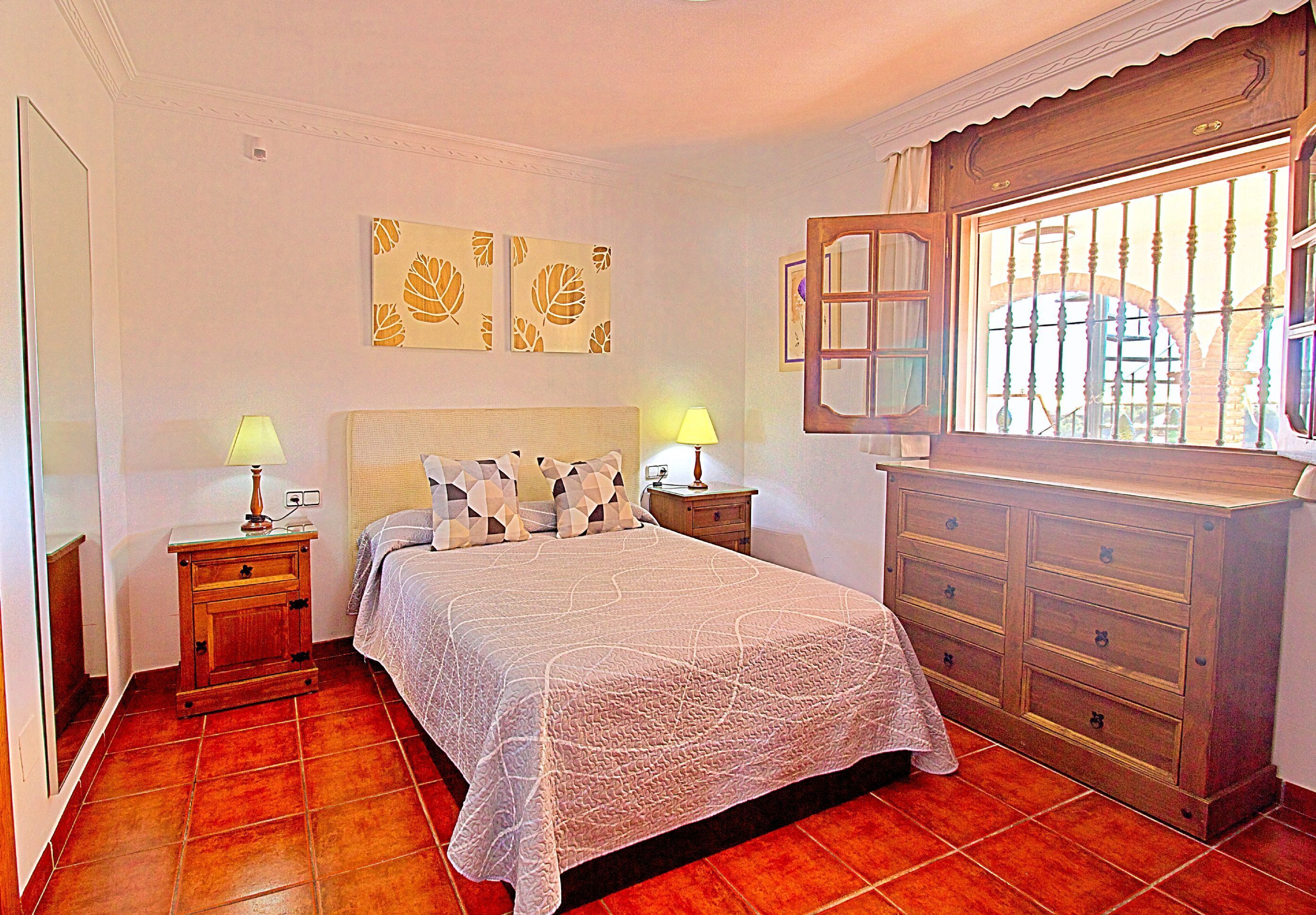 A bedroom with a bed, two nightstands with lamps, a dresser, and a window with bars.