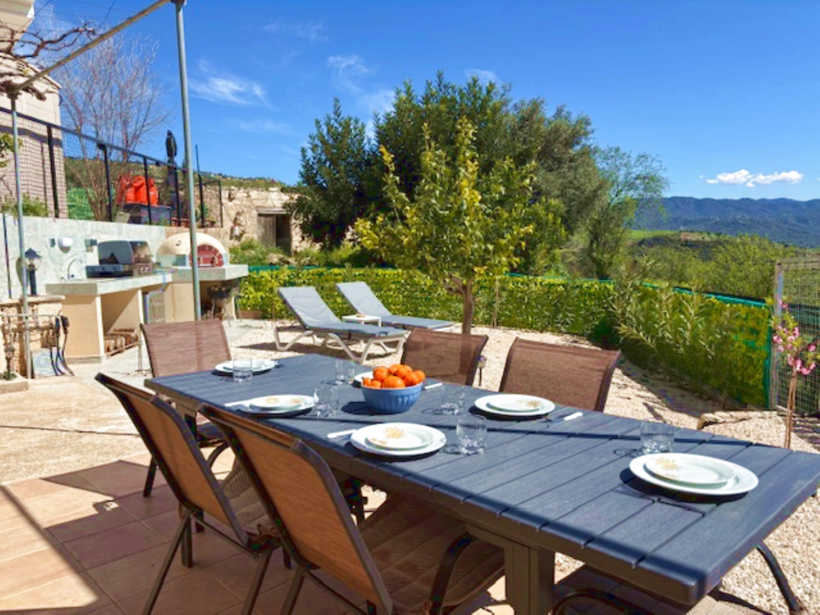 Un espace extérieur aménagé pour prendre les repas, avec une table dressée pour deux personnes, des chaises et un saladier rempli de fruits.