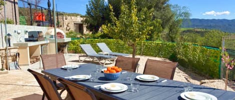 Un espace extérieur aménagé pour prendre les repas, avec une table dressée pour deux personnes, des chaises et un saladier rempli de fruits.