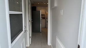 Un couloir étroit avec une porte sur la gauche, un spot encastré au plafond et, au fond, un espace cuisine.