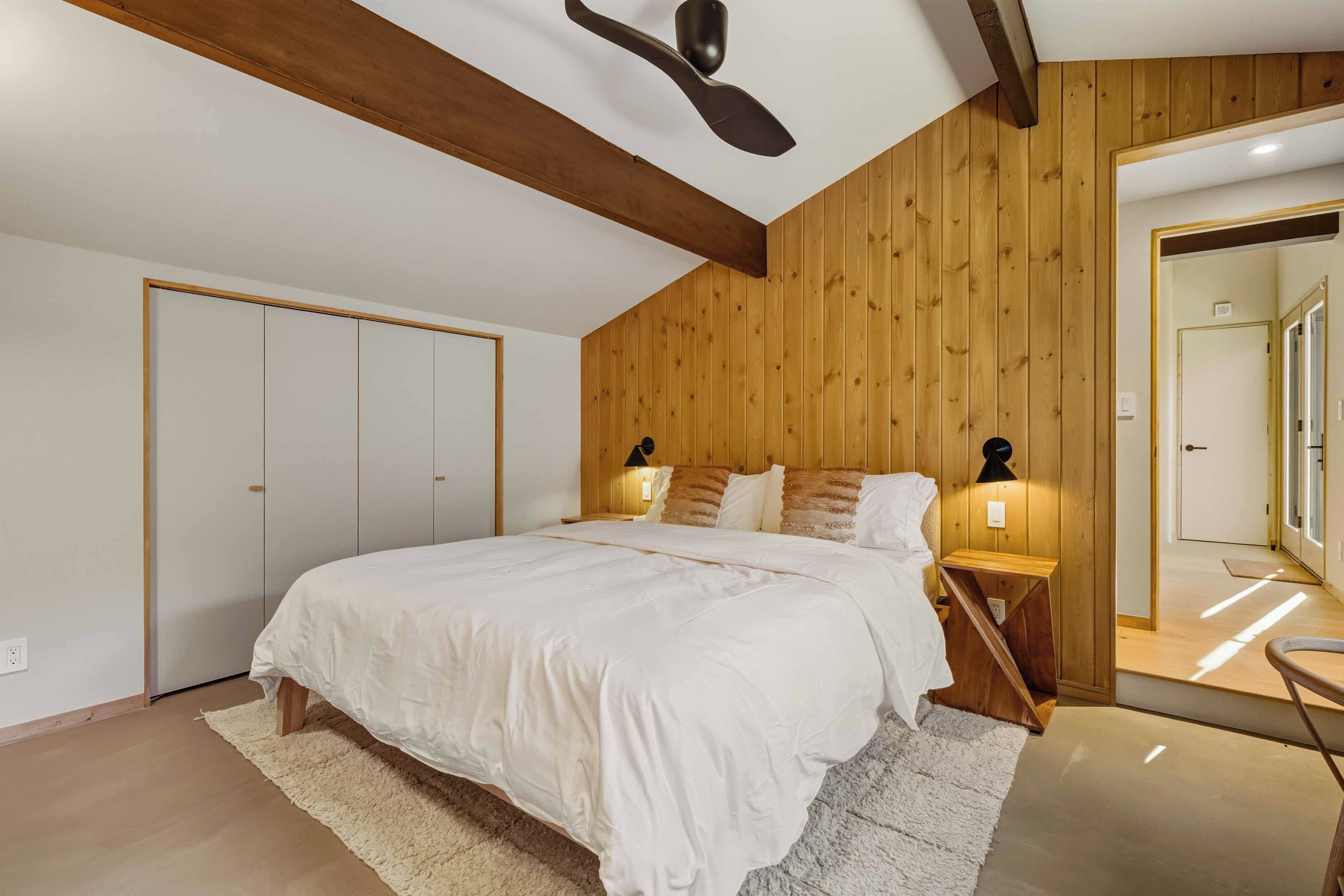 A bedroom with a wooden ceiling, a bed with white bedding, two bedside tables with lamps, and a large mirror on the wall.