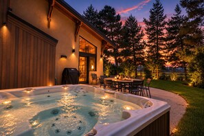 A hot tub in a backyard with a patio area and a building with a wooden exterior. - The Sawtooth Modern | Designer Build | Hot Tub | River Access (Ketchum)