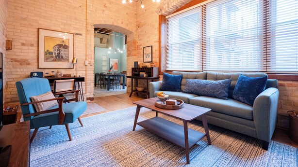 Ein gemütliches Wohnzimmer mit Backsteinwand, einem blauen Sessel, einer Couch, einem Couchtisch und einem großen Fenster mit Jalousien.