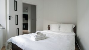 Una cama bien tendida con sábanas blancas y una toalla doblada, un cabecero de madera y una pared blanca.