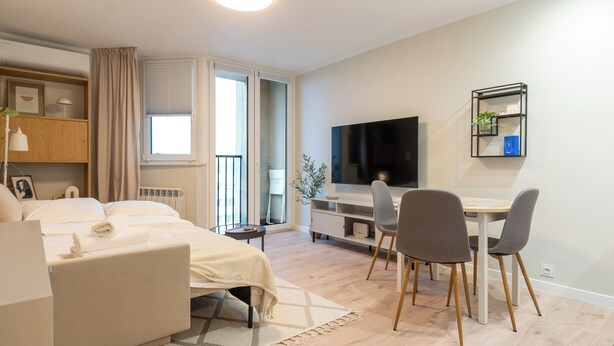 Habitación de hotel moderna con cama, escritorio, dos sillas, televisor y balcón con puerta corrediza.