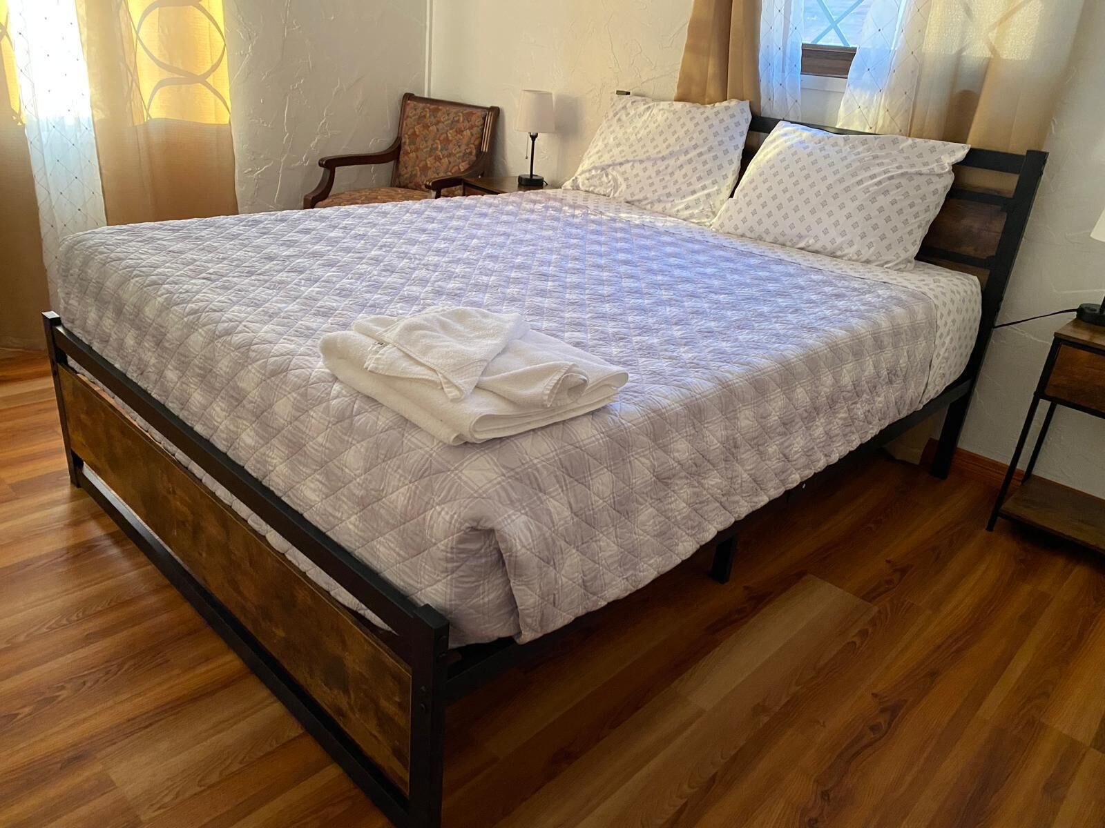 A neatly made bed with a quilted bedspread and folded towels, a wooden nightstand with a lamp, and a chair in a room with wooden flooring and a window with sheer curtains.