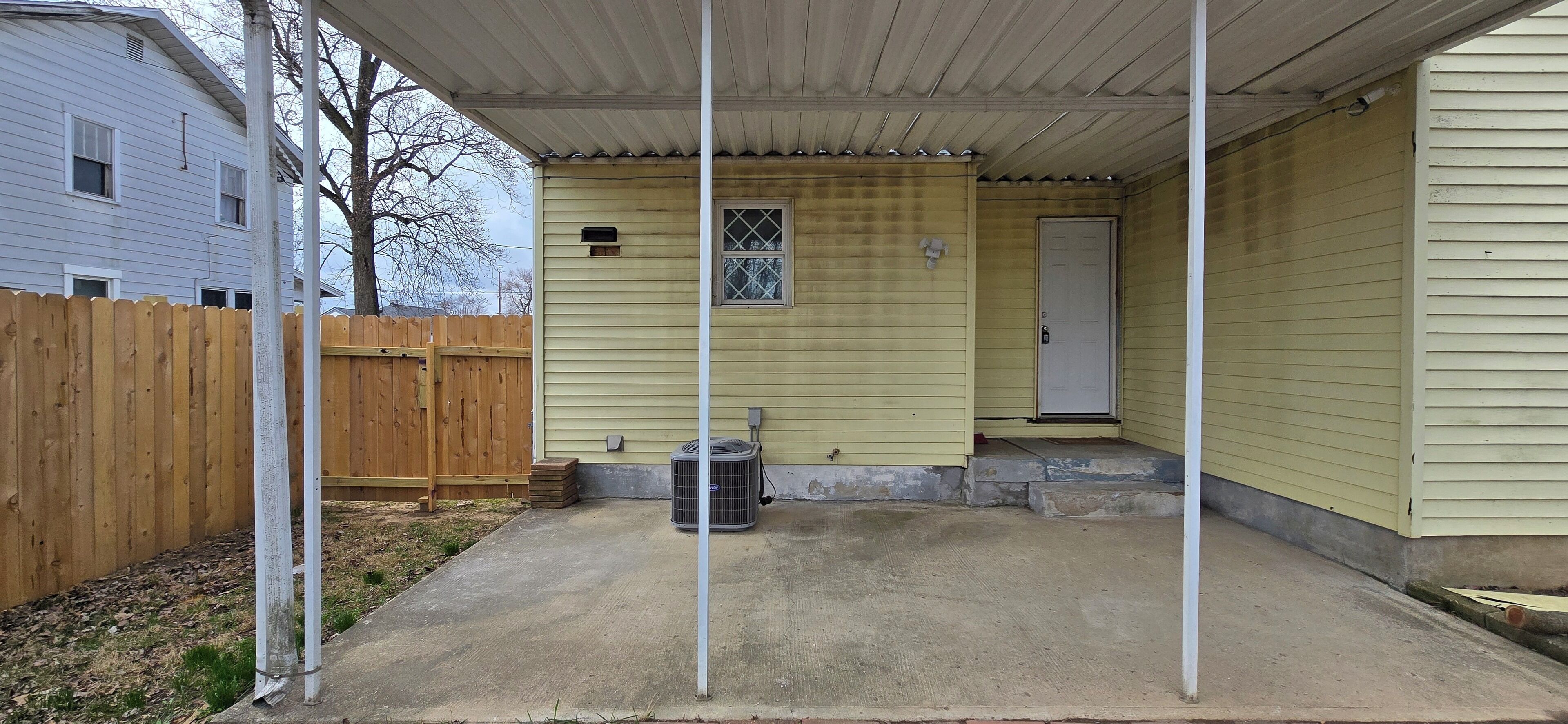 A covered patio area with a concrete floor, a wooden fence, and a small air conditioning unit.