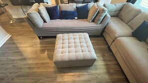 A living room with a beige sectional sofa, a matching ottoman, and a wooden floor. - Masters 2026 Rental Home w/Additional Loft in Grovetown, GA! (Grovetown)