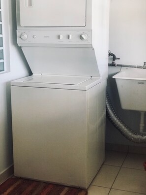 A compact, white front-loading washing machine in a small laundry room with a visible hose and a sink. - Apartments Pepe's View - C (Aguada)