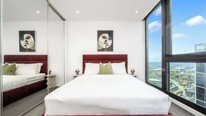 Une chambre d’hôtel moderne avec un grand lit, une tête de lit rouge et une vue sur la ville grâce à de grandes fenêtres.