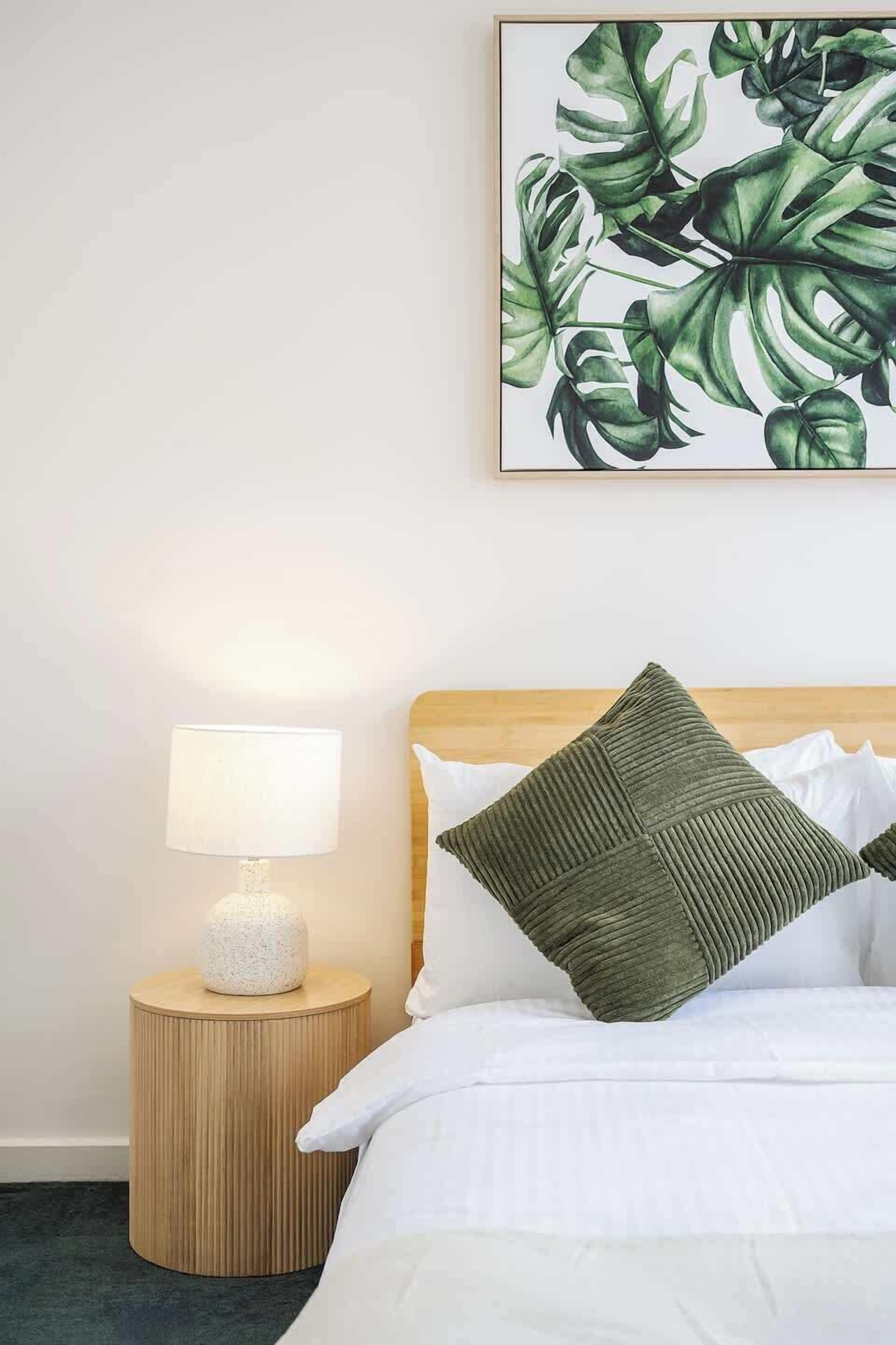 A neatly made bed with white bedding and a green pillow, a round wooden nightstand with a lamp, and a framed botanical print on the wall.