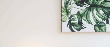 A neatly made bed with white bedding and a green pillow, a round wooden nightstand with a lamp, and a framed botanical print on the wall.