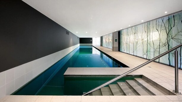 Une piscine intérieure moderne offrant une vue dégagée sur le bassin, un escalier menant au bord de la piscine et un mur de verre orné d’un motif d’arbre.