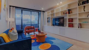 Un salon moderne avec un canapé bleu, un fauteuil rouge, une table basse ronde et un téléviseur posé sur une étagère blanche.
