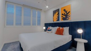 Une chambre à coucher comprenant un lit, deux lampes de chevet, une fenêtre avec des stores et deux œuvres d’art murales.