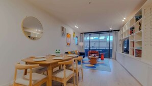 Une pièce moderne et bien éclairée, avec une table à manger en bois dressée pour quatre personnes, un grand miroir rond accroché au mur et une vaste fenêtre donnant sur le paysage urbain.