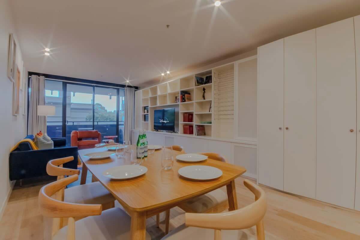 A modern dining area with a wooden table set for four, surrounded by wooden chairs. There is a TV on a shelf, and a large window providing a view outside.