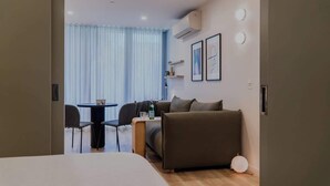 A modern hotel room with a bed, a sofa, a small table, and a chair. There is a window with curtains, an air conditioning unit on the wall, and framed pictures. - Scandinavian Suite, Lap Pool, Big Gym, Melbourne (South Yarra)
