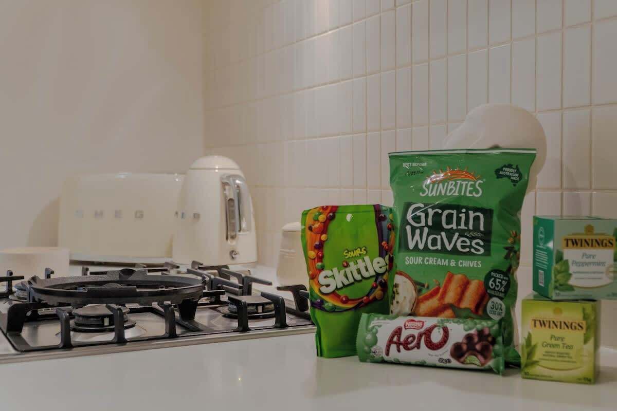 Eine Küchenarbeitsplatte mit einem Gasherd, einer Kaffeemaschine und verschiedenen Lebensmitteln, darunter Skittles, Sunbites Grain Waves, Aero und Twinings Pure Green Tea.