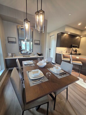 A modern dining area with a wooden table set for four, a kitchen with dark wooden cabinets and a microwave, and pendant lights above the table.