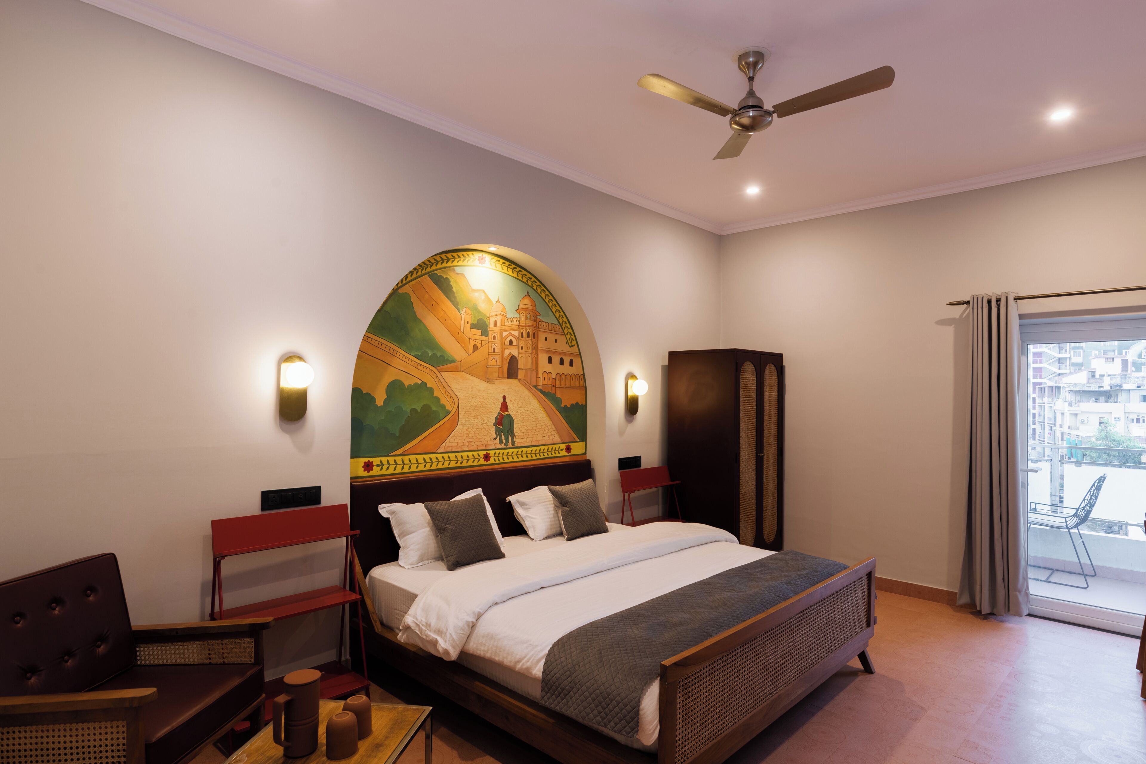A hotel room with a bed, a ceiling fan, a wall mural, a red chair, and a view of the city through a window.