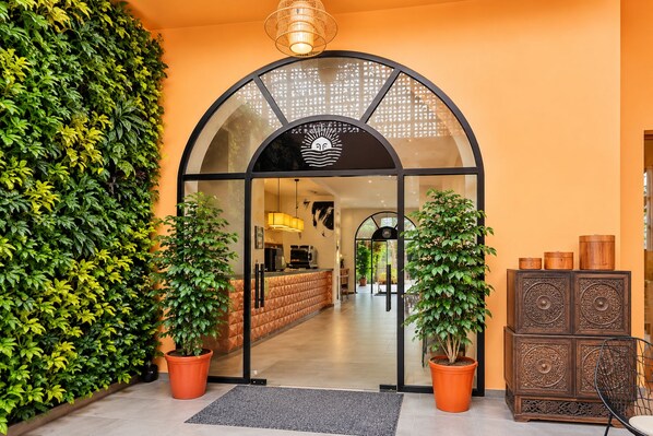 Entrada de un hotel con puerta de vidrio, plantas en macetas, un mostrador de recepción y un aparador decorativo.
