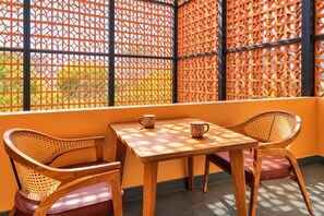 A room with a wooden table and two wicker chairs, a window with a lattice screen, and a view of greenery outside. - Nidra Niwas (Jaipur)