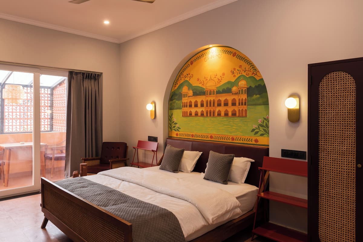 A hotel room with a bed, a painting of a building on the wall, a door, and a view of the outside.
