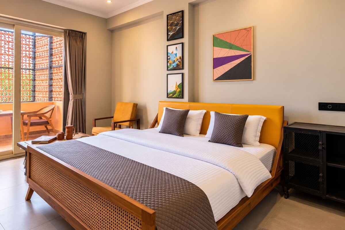 A hotel room with a large bed, a dining table, chairs, a balcony with a view, and artwork on the walls.