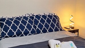 A neatly made bed with a blue and white patterned bedspread, a set of folded towels, and a wall-mounted picture of a sunset over the ocean. - Spacious 5-Bd Tampa Retreat with Dual Jacuzzis. Close to Top Attractions. (Tampa)