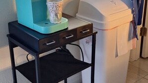A coffee maker on a black metal table next to a white trash bin and a window with a view of trees. - Spacious 5-Bd Tampa Retreat with Dual Jacuzzis. Close to Top Attractions. (Tampa)
