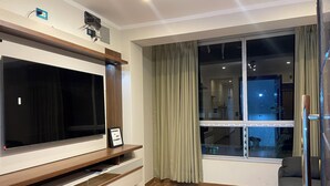 A modern living room with a large flat-screen TV mounted on a wooden console, a glass door leading to a balcony, and a view of the outside.