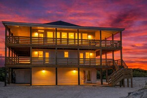 Une maison de plage en bois à deux étages, dotée de balcons, d’un porche couvert et d’un escalier menant à l’étage supérieur.