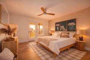 Une chambre à coucher comprenant un lit, des tables de chevet, un ventilateur de plafond, une grande fenêtre et une vue sur une maison.