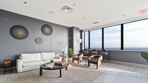 Quarto de hotel moderno com sofá branco, mesa de centro, escrivaninha e cadeiras. Janelas amplas oferecem vista para a cidade.