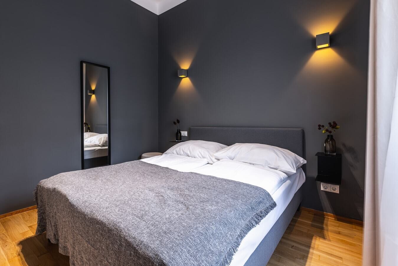 A modern bedroom with a large bed, a bedside table, a mirror, and two wall-mounted lights.