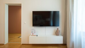 Un salon moderne avec un téléviseur à écran plat fixé à un meuble blanc, une petite table sur laquelle est posé un vase, et une porte donnant sur une autre pièce.