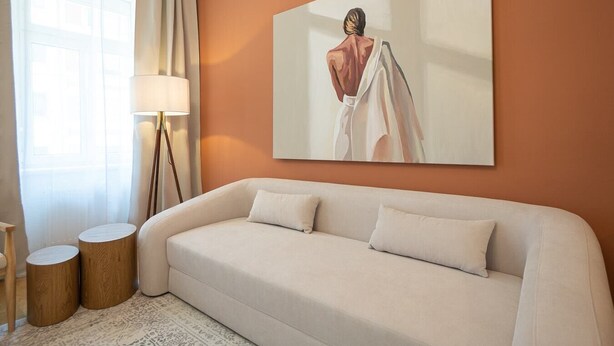 Un salon moderne avec un canapé beige, deux petites tables d’appoint rondes en bois, une lampe sur pied et un grand tableau accroché au mur.
