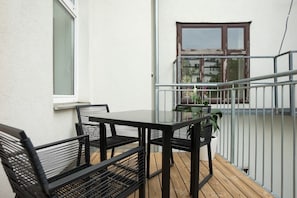 Een balkon met een glazen tafel en zwarte metalen stoelen, een witte muur en een raam met bruin kozijn.