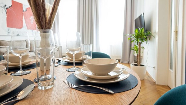 Mesa de comedor dispuesta con platos, tazones, vasos y cubiertos, con un florero de plantas secas en el centro.