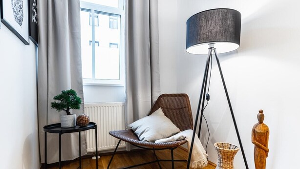 A cozy room with a wooden floor, a standing floor lamp, a chair with a cushion, a small table with a potted plant, and a decorative wooden sculpture. - Precious apartment in calm district area (Wien)