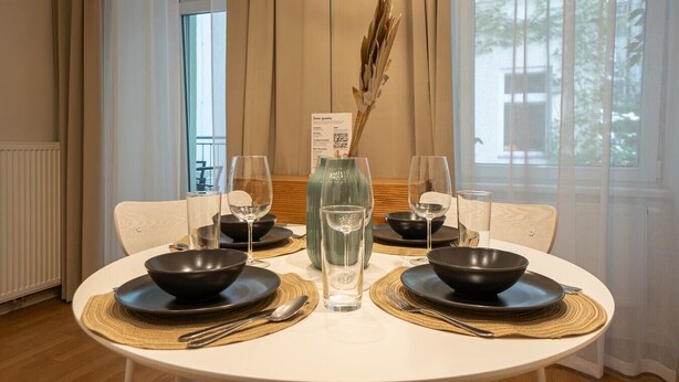 Une table à manger dressée pour quatre personnes, avec des bols noirs, des verres à vin et un vase contenant des fleurs séchées.