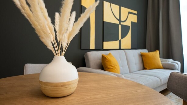 Una sala moderna con una mesa de comedor de madera, un florero blanco con plumas de pampa, un sofá gris con cojines amarillos y un cuadro abstracto negro y dorado en la pared.
