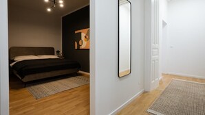 Un dormitorio con una cama, una mesita de noche, una lámpara moderna, un espejo en la pared y una puerta que da a otra habitación.